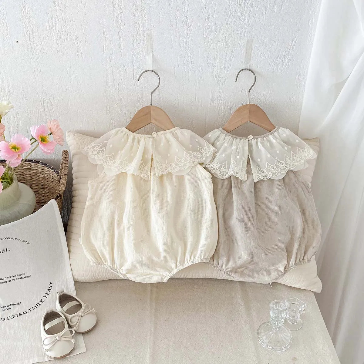 A beige baby girl sleeveless jumpsuit with a lace collar displayed on a white bed with natural light.