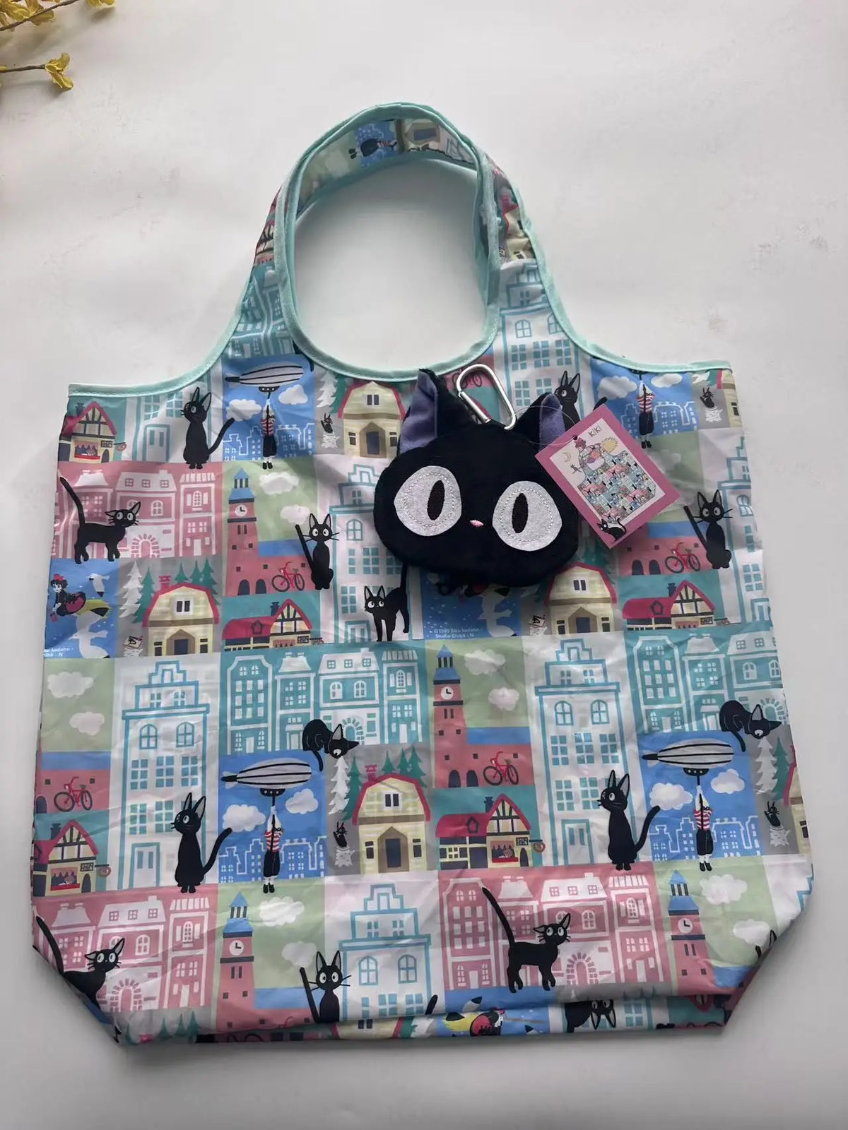 A patterned fabric tote bag featuring a cute black cat design on a light blue and pink background.