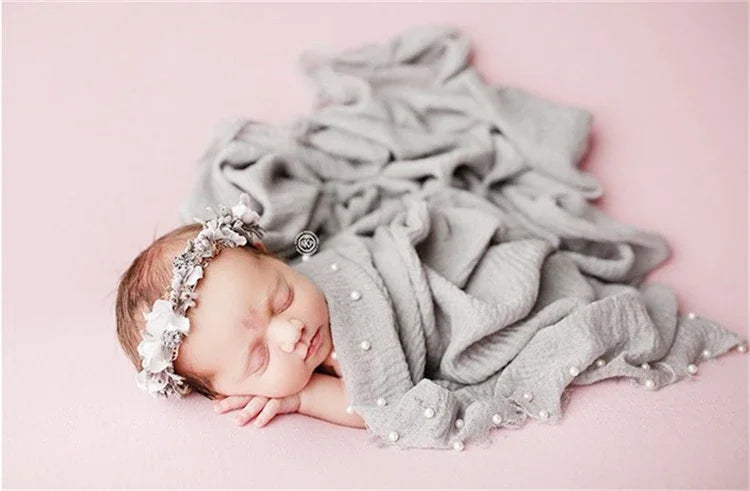 A newborn baby sleeping peacefully wrapped in a gray knit wrap with a pearl headband.