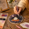 A hand paints a Halloween diamond painting ornament featuring a white ghost on an orange pumpkin amidst scattered glitter and