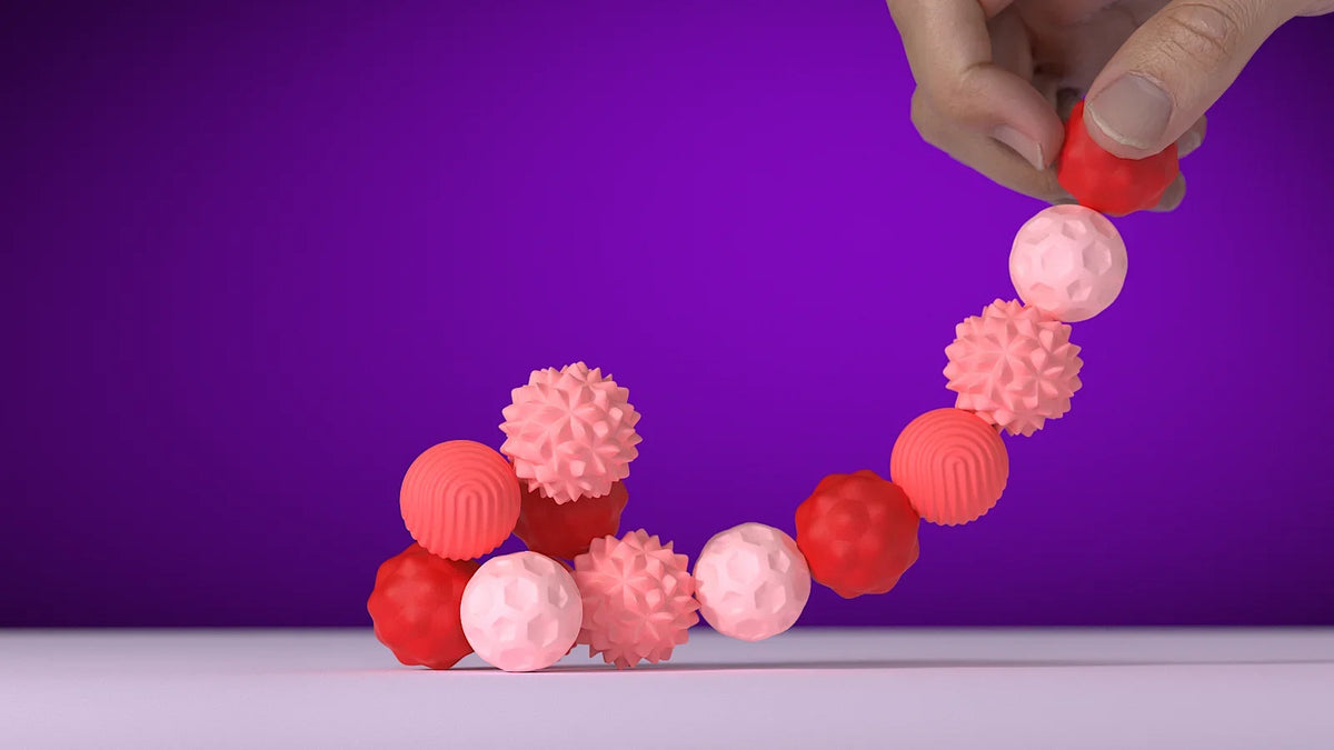 A hand is holding a string of pink and red soft sensory silicone massage balls against a purple background.