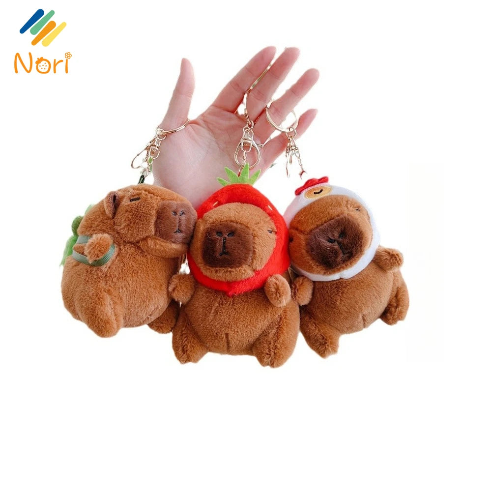 A hand holds three adorable capybara plush keychain charms with gold-colored chains.
