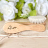 A light wooden baby brush and comb set with the name Alice engraved on the handle, resting on a beige cloth.