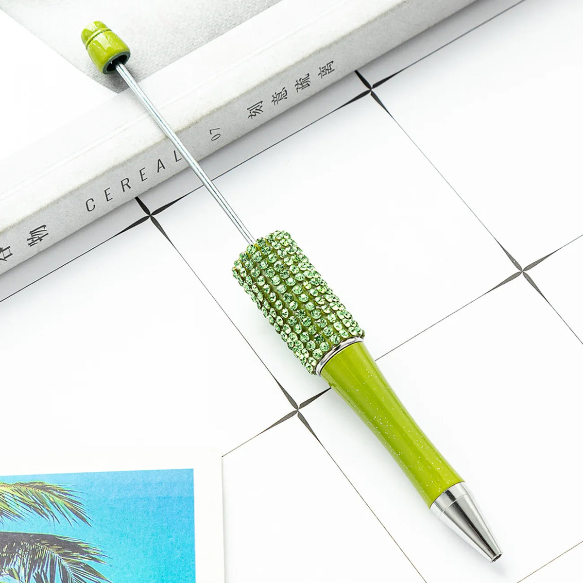 A green beaded ballpoint pen craft kit with a metal tip rests on a white surface next to a book.