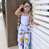 A young girl wearing a white suspender top and colorful long pants with sunglasses standing next to a white wall.