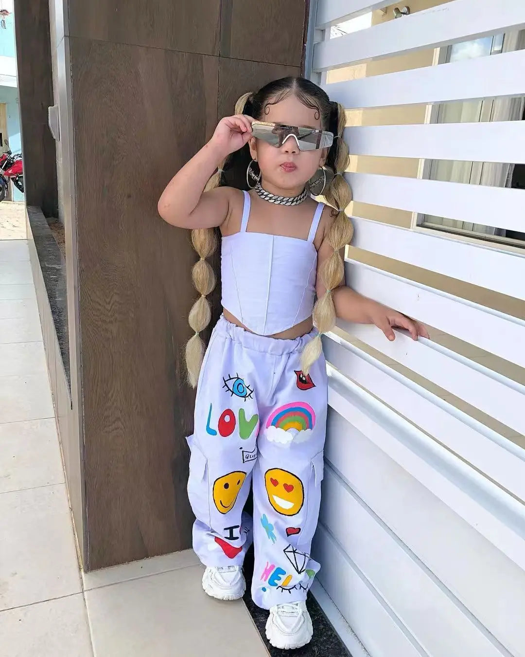 A young girl wearing a white suspender top and colorful long pants with sunglasses standing next to a white wall.