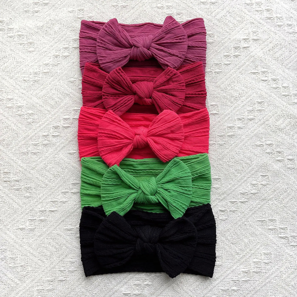 A stack of five colorful fabric bow baby headbands in shades of pink, red, coral, green, and black.