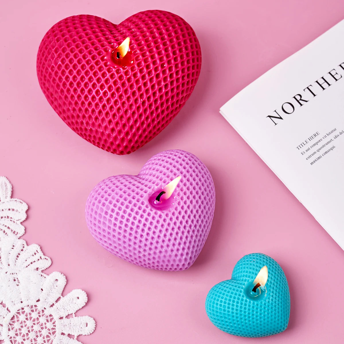 A set of three silicone heart candle molds in pink, purple, and teal resting on a pink surface.