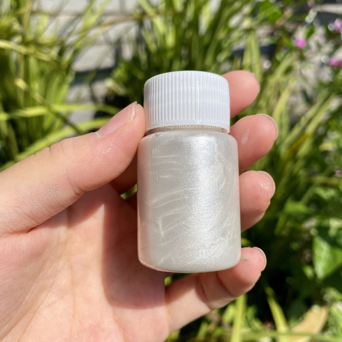 A hand holds a small glass bottle of metallic gold acrylic paint for crafts against a blurred green outdoor background.