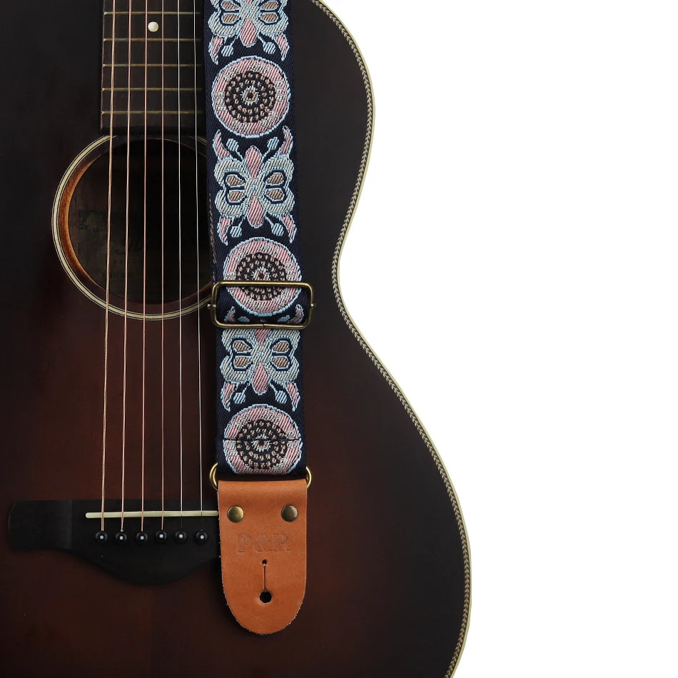 An embroidered guitar strap featuring blue and white patterns with leather ends attached to a dark acoustic guitar neck.
