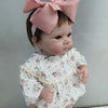A lifelike 18-inch reborn baby doll with hand-painted details wearing a white floral outfit and a pink bow.