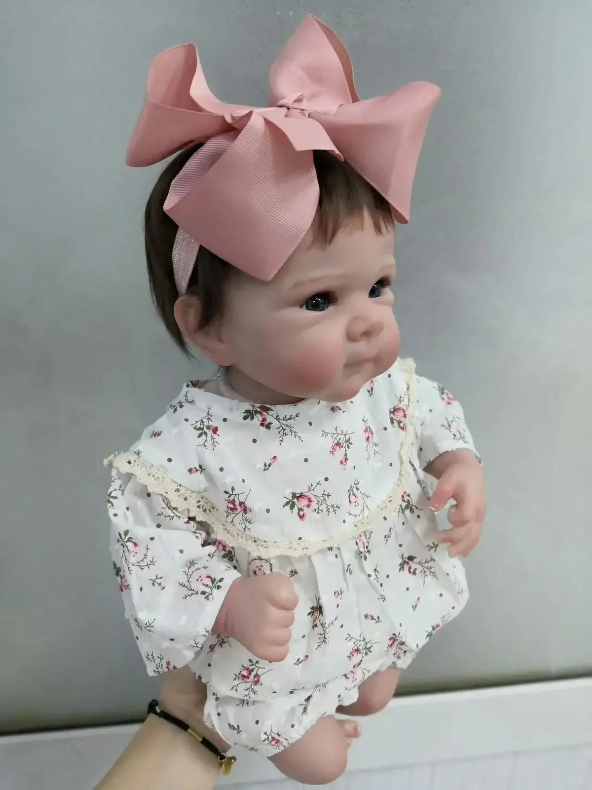 A lifelike 18-inch reborn baby doll with hand-painted details wearing a white floral outfit and a pink bow.