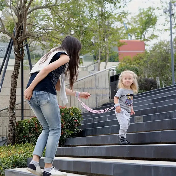 A young girl is walking down outdoor stairs while being led by an adult using a pink anti-lost wrist harness.