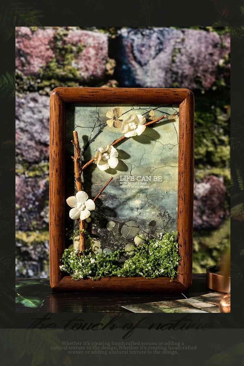 A framed nature scene featuring dried branches, white flowers, and moss on a textured background.