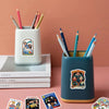 A teal ceramic mug filled with colored pencils sits next to a stack of books and a sticker pack.