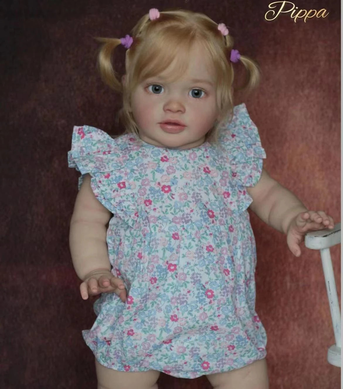 A realistic reborn baby doll kit named Pippa with blonde hair and pink hair accessories wearing a floral dress.