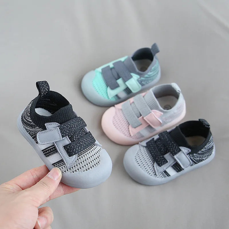 A hand holds a pair of breathable mesh toddler walking shoes in gray and pink tones.