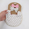 A set of three pink and white floral baby flower bibs and a matching bow headband displayed on brown cardstock.