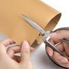 Hands are using a pair of scissors to cut a self-adhesive PU leather repair patch from a tan material.