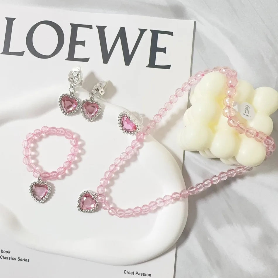 A pink pearl and rhinestone jewelry set featuring earrings, a necklace, and a charm on a white ceramic dish.