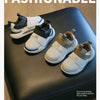 A pair of light gray and white kids' everyday sneakers with easy velcro straps resting on a dark surface.