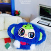 A blue fuzzy character pendant with large eyes and green accents is displayed on a white surface next to a laptop.