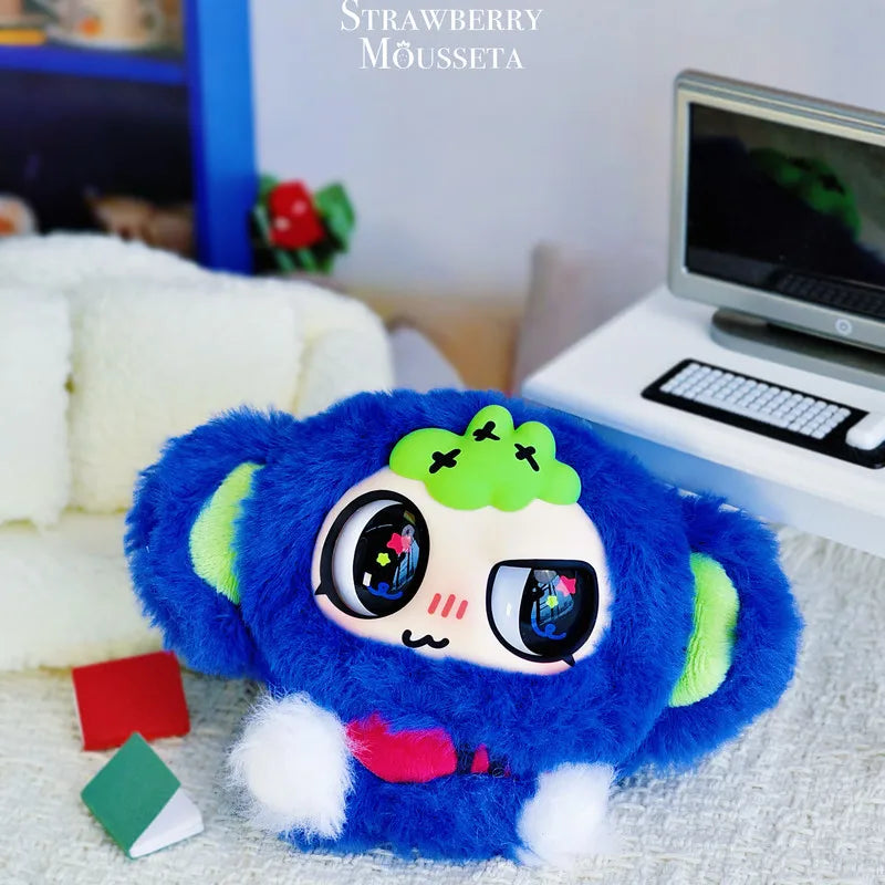 A blue fuzzy character pendant with large eyes and green accents is displayed on a white surface next to a laptop.