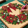 A hand is decorating a Christmas wreath made of red and green beads, pearls, and gold accents on a cardboard surface.