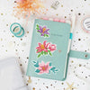 A teal notebook featuring floral designs and stickers, surrounded by glitter and star confetti.