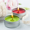 A small silver tin candle with a green flame sits on a white wooden surface next to pink flowers.