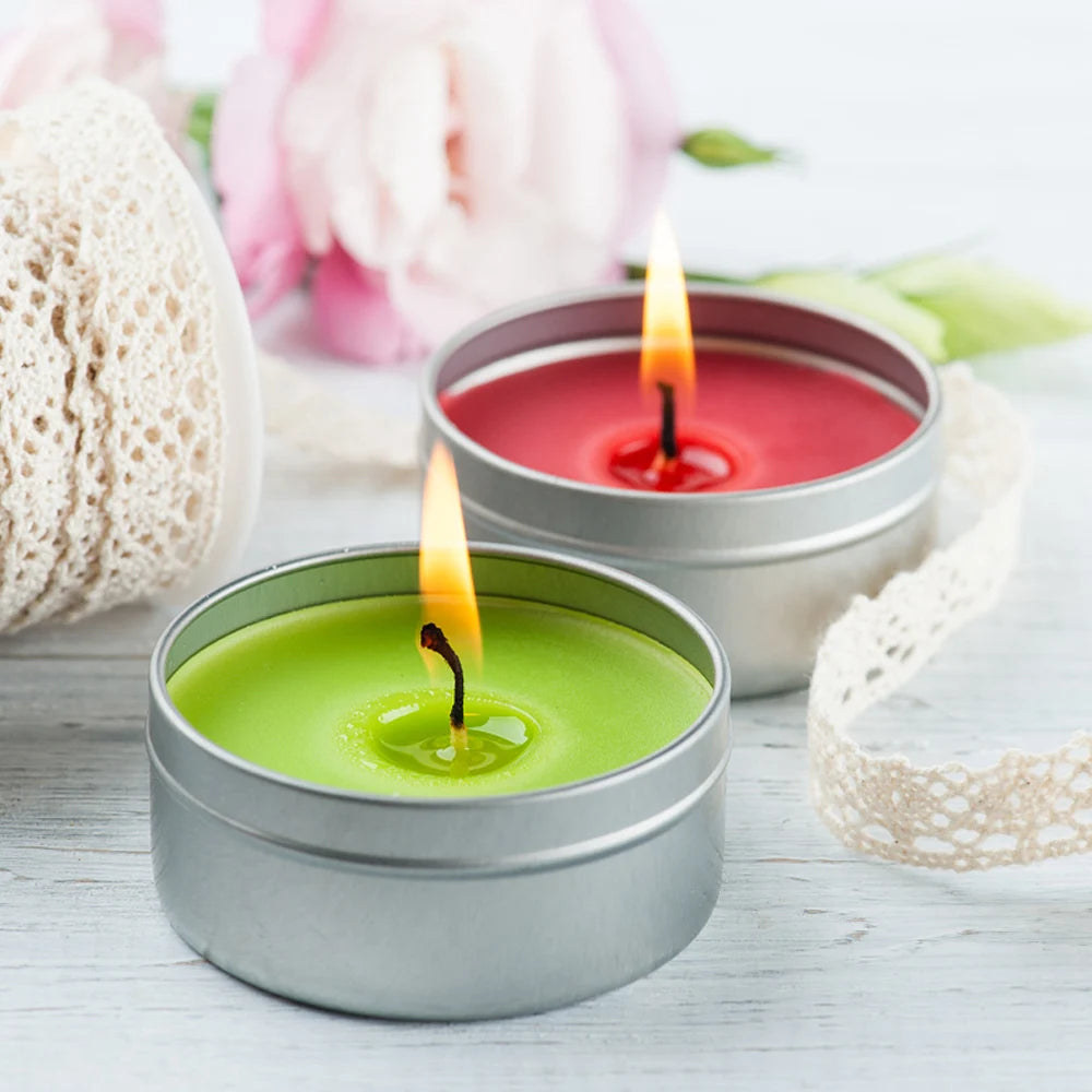 A small silver tin candle with a green flame sits on a white wooden surface next to pink flowers.