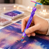 A hand holds a purple diamond painting pen with an interchangeable resin tip, writing on a colorful canvas.