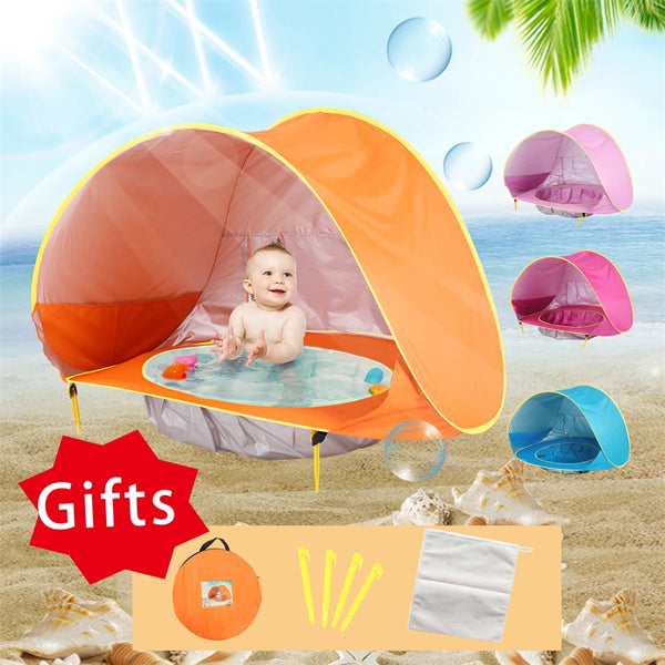 An orange portable baby beach tent is open, with a baby inside, set up on a sandy beach with water and tropical foliage.