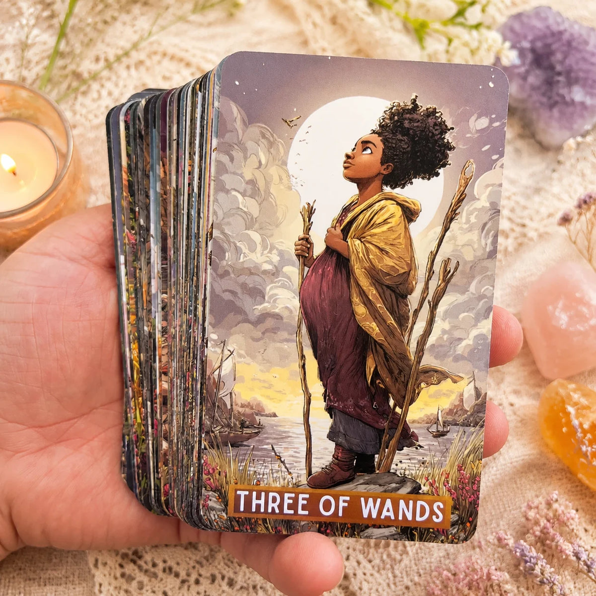 A hand holds a tarot card depicting a woman with curly hair standing by water under a large moon, labeled Three of Wands.