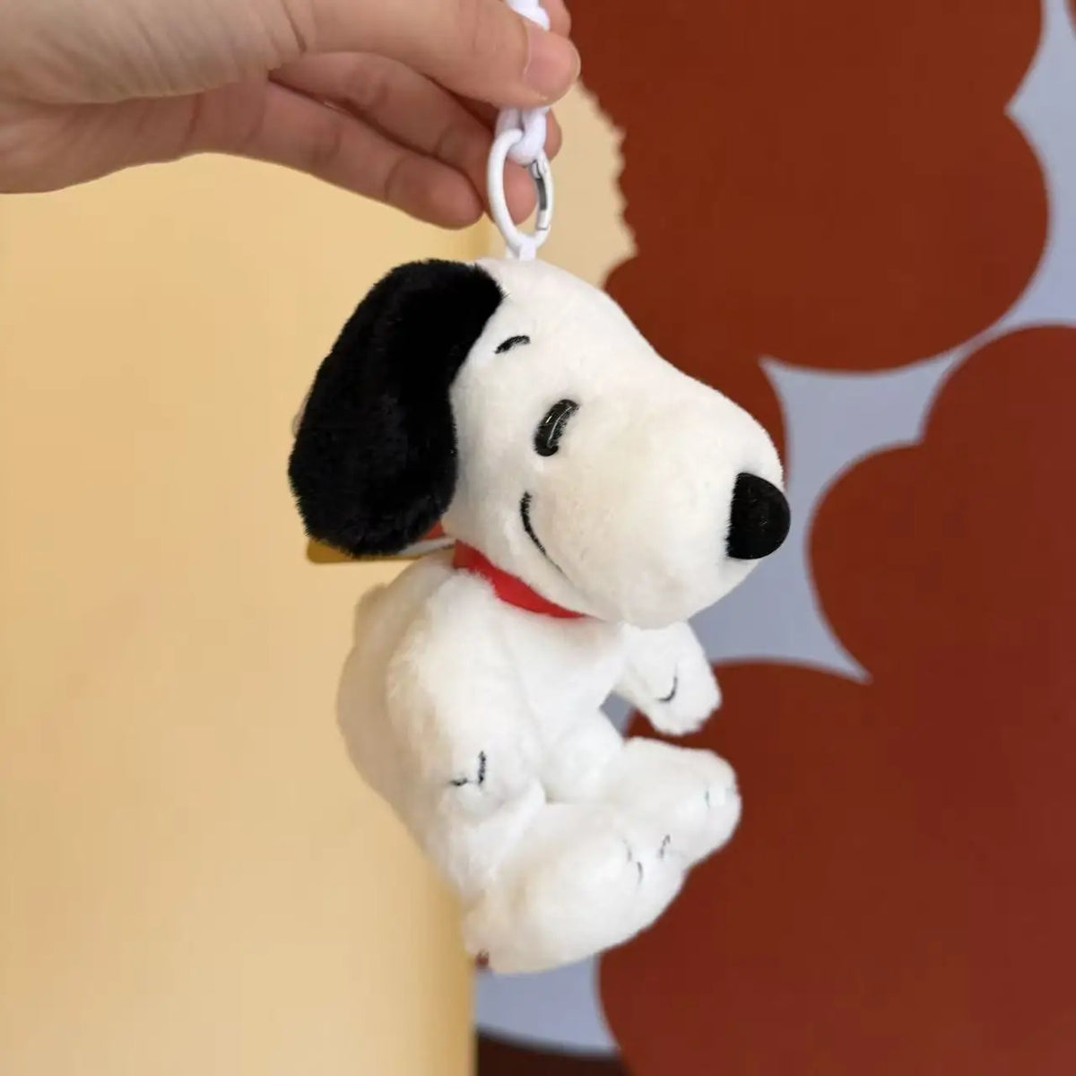 A white Snoopy plush keychain charm with black ears and a red collar is being held by a hand.