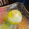 A translucent yellow fruit squishy stress relief ball resting on a reflective glass plate.