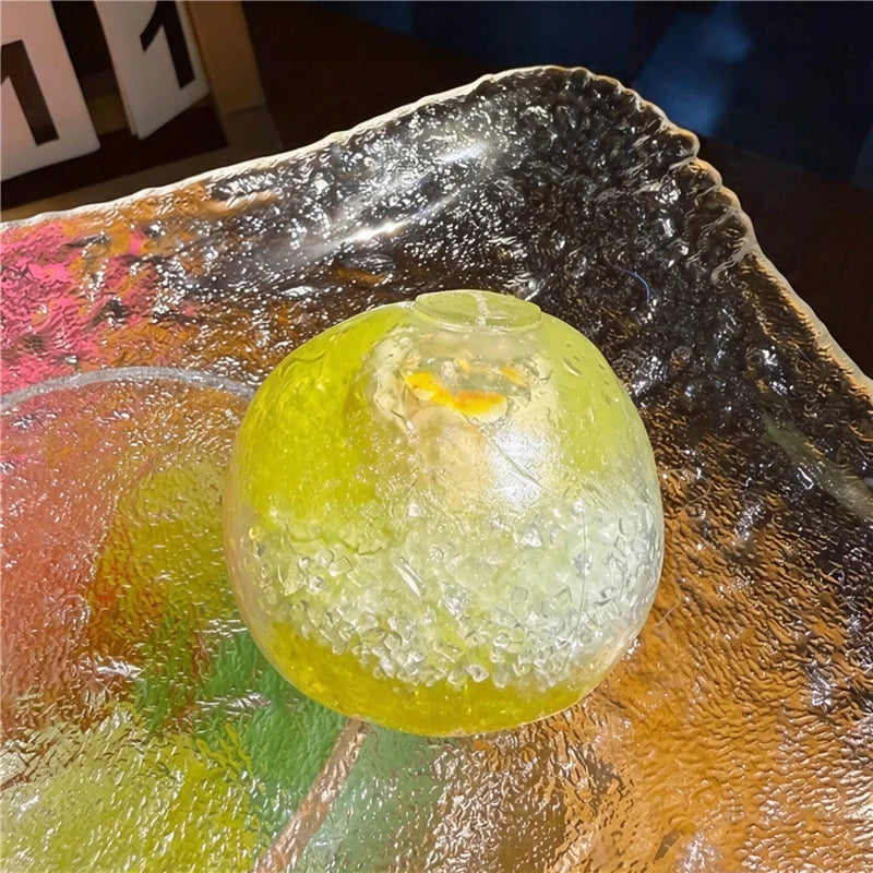 A translucent yellow fruit squishy stress relief ball resting on a reflective glass plate.