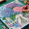A hand paints a floral design on a canvas using pink and blue pearls and beads within a wooden frame.