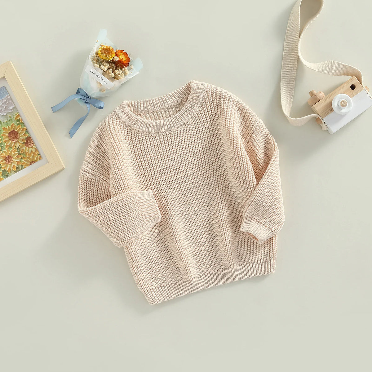 A beige knit pullover sweater for kids with long sleeves, displayed on a light surface next to a small potted plant and a lig