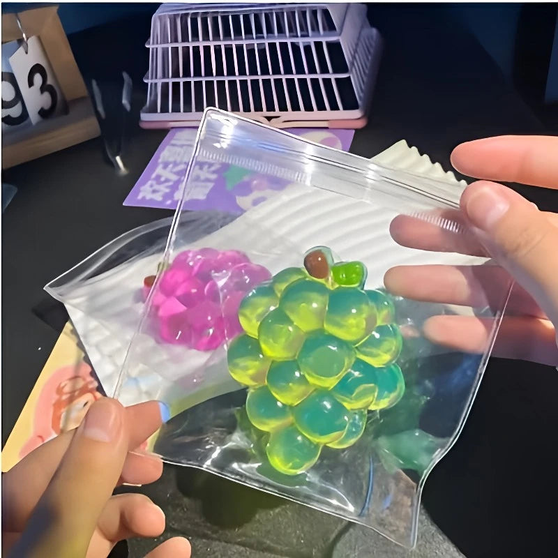 A hand holds a clear plastic bag containing a cluster of translucent green and yellow squishy grape fidget toys.
