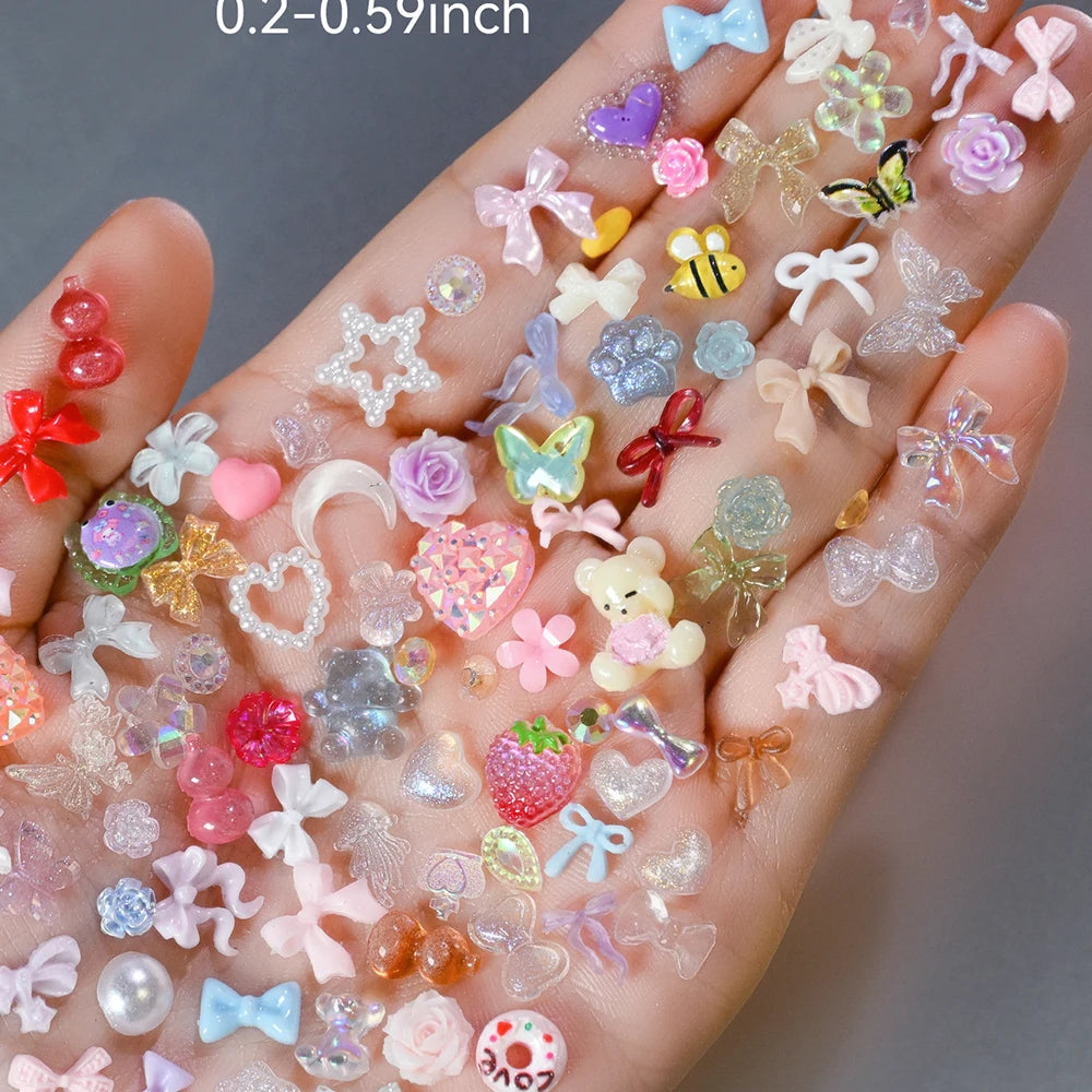 A hand displays a variety of multicolor resin nail charms including hearts, flowers, butterflies, and various glitter accents