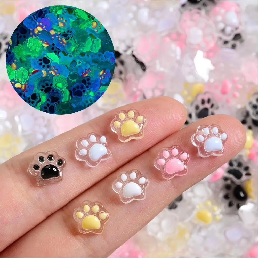 A hand displays various 3D luminous cat paw nail art charms in pastel colors and a green circular design.