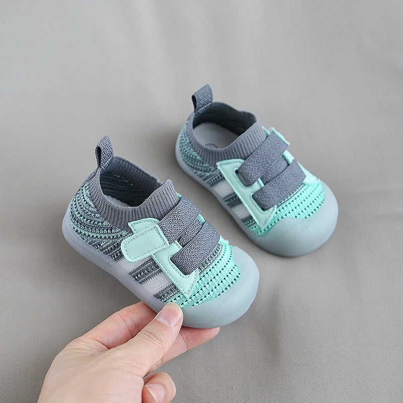 A pair of light mint green and gray breathable mesh toddler walking shoes are being held by a hand.