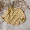 A light yellow long sleeve cotton tee is laid on a white surface next to a knitted beige fabric and dried plant stems.