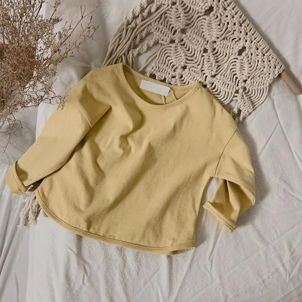 A light yellow long sleeve cotton tee is laid on a white surface next to a knitted beige fabric and dried plant stems.