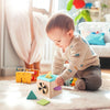 A baby is playing with a wooden Montessori magic tissue box toy and small green plastic shapes on a rug.