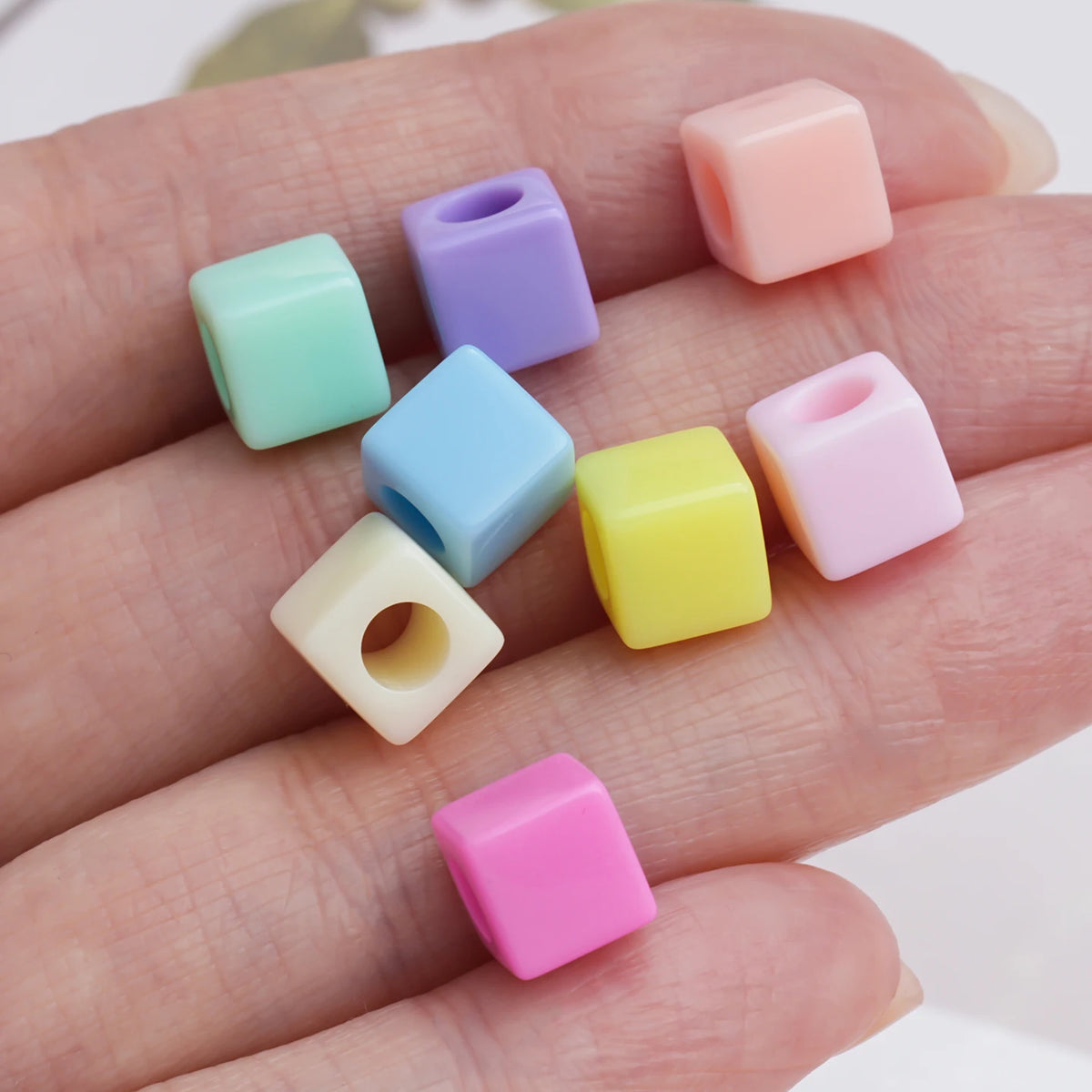 A hand holds several square acrylic spacer beads in various pastel colors including pink, purple, mint green, and yellow.