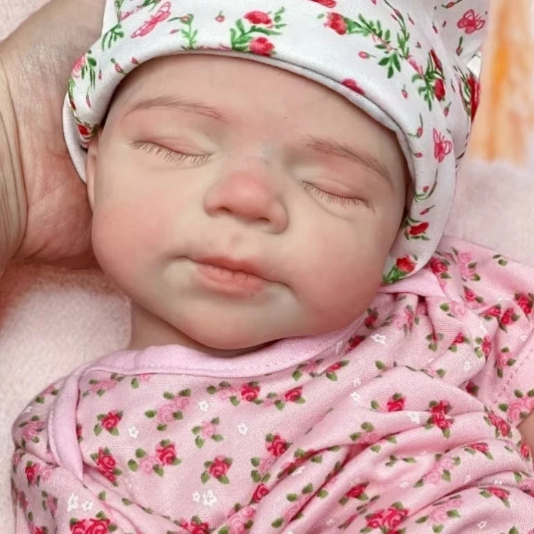 A close-up photo of a lifelike silicone reborn baby doll with pink floral clothing, sleeping peacefully.