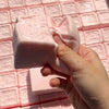 A hand holds a pink strawberry milk squishy stress relief toy against a pink grid background.