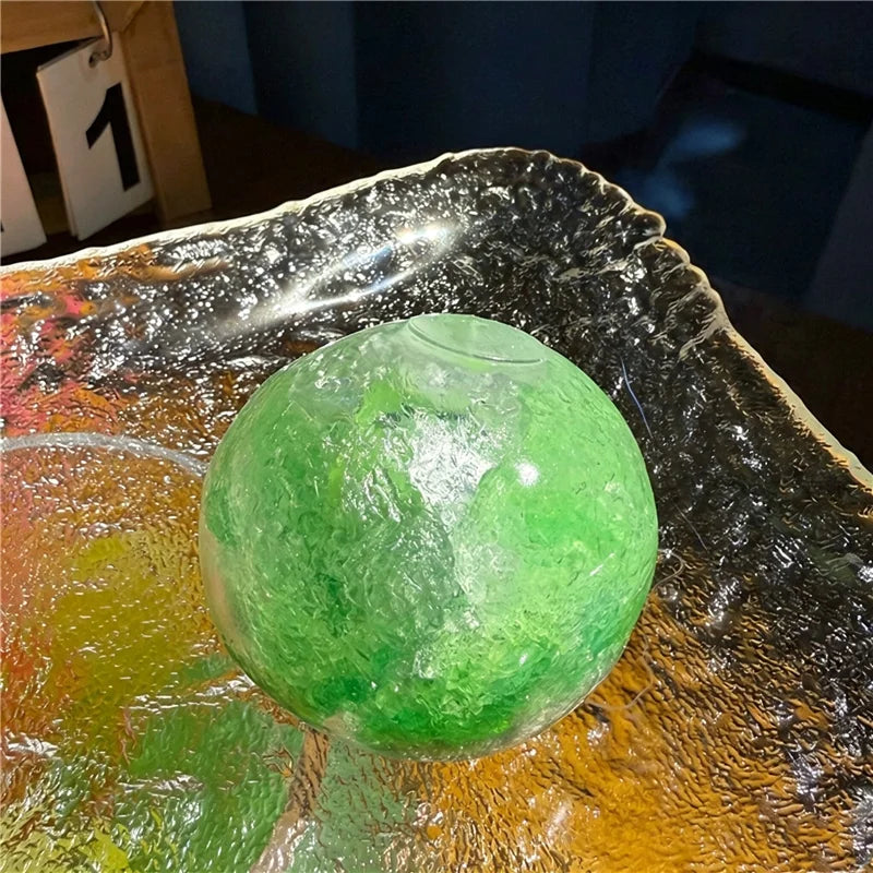 A translucent green fruit squishy stress relief ball resting in a reflective, textured dish.